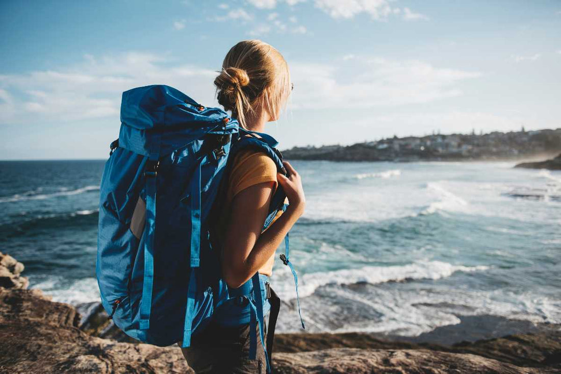 Backpacker with blue pack looking over rocky coastline and waves, adventure.