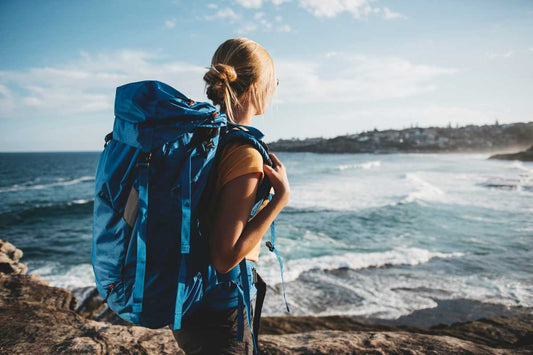 Backpacker with blue pack looking over rocky coastline and waves, adventure.