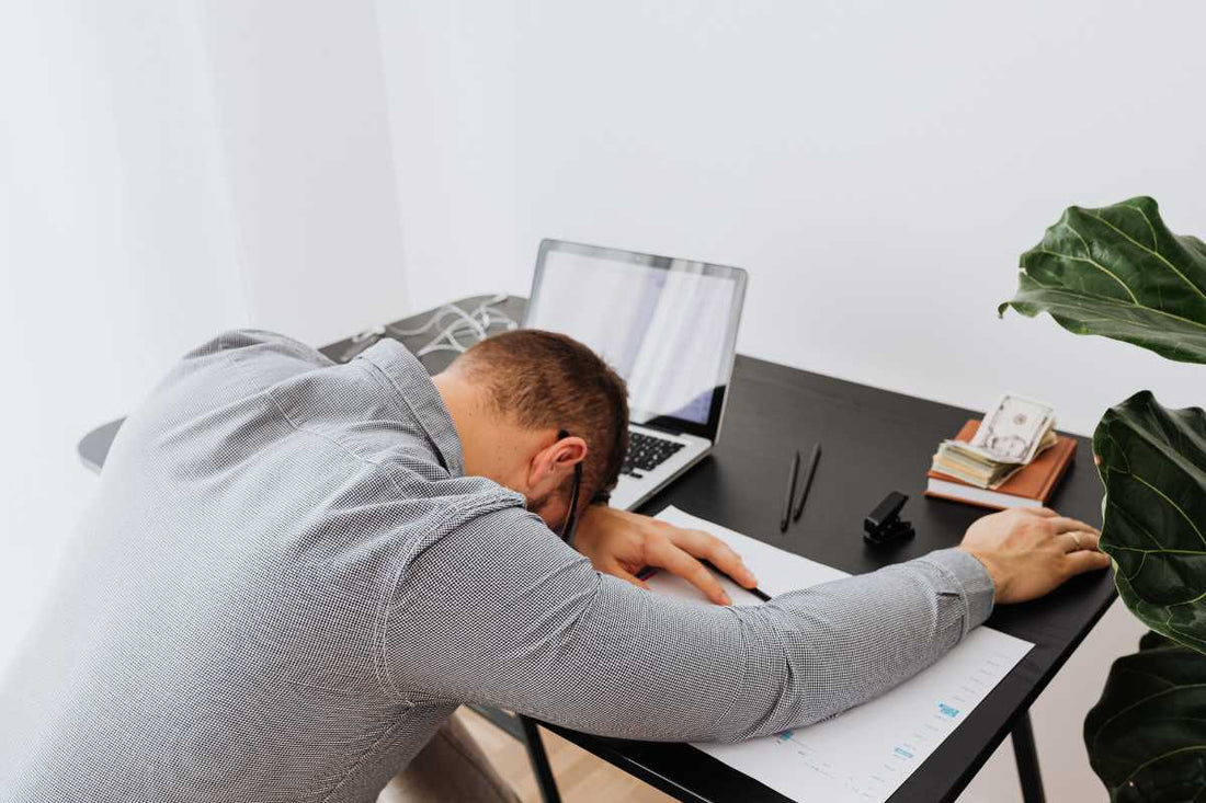 Man resting face-down on a desk beside a laptop and paperwork, fatigue
